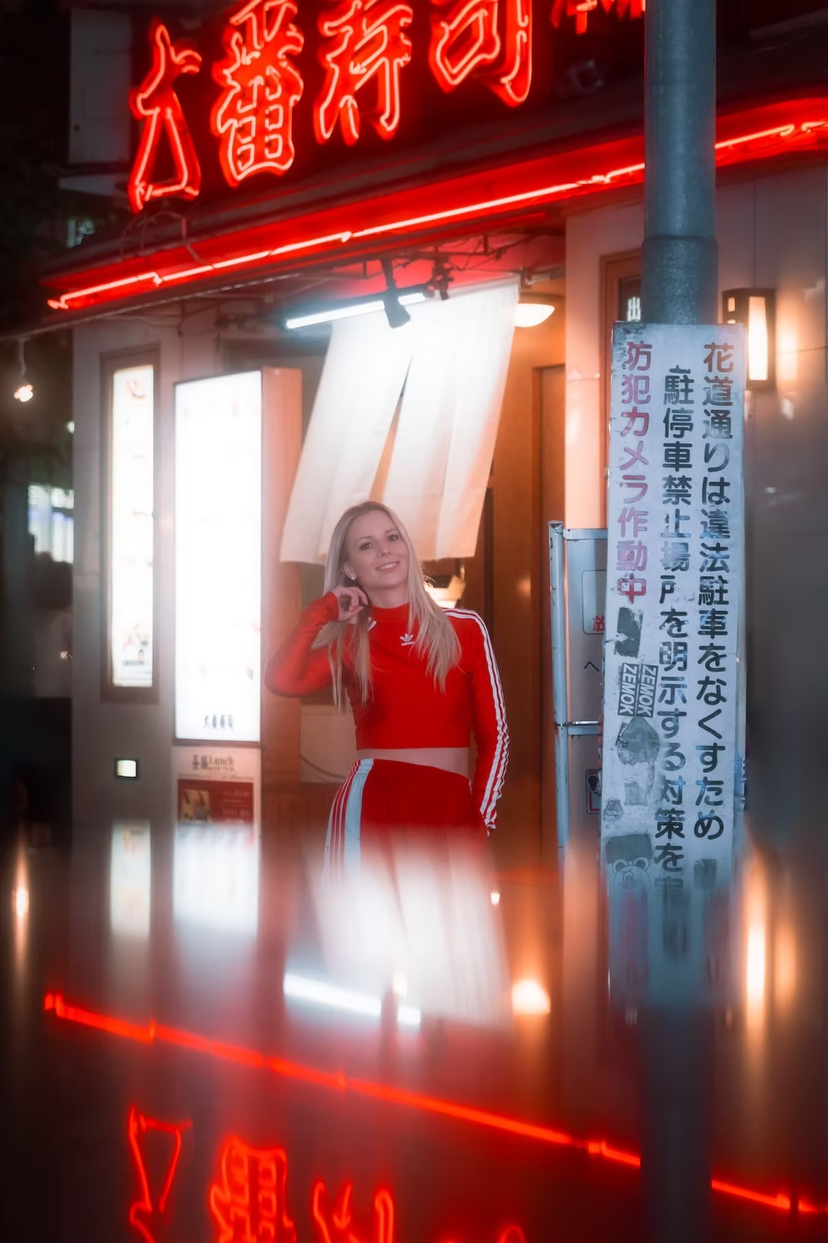 A woman wearing a red tracksuit stands in front of a brightly lit sign in a Tokyo alley, with neon lights illuminating her surroundings.