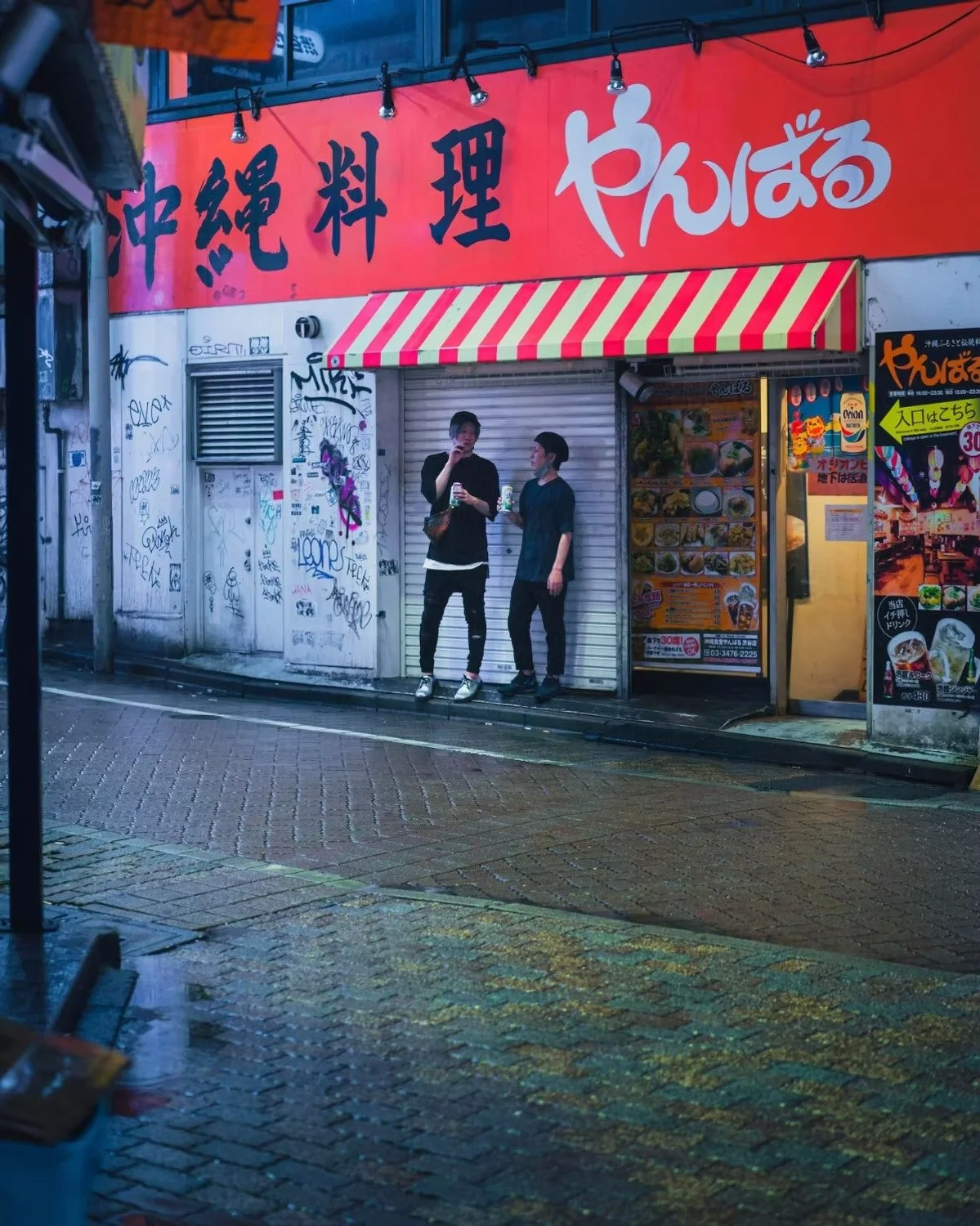 Two young men are standing on a rainy street in front of a Japanese restaurant with red signage and a red striped awning. They are holding drinks and talking to each other, with graffiti on the wall behind them. Shot by Tokyo street photographer Deni