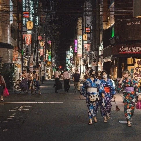 A lively Japanese street at night with women in colorful yukatas walking together, surrounded by neon signs, lanterns, and bustling pedestrians.