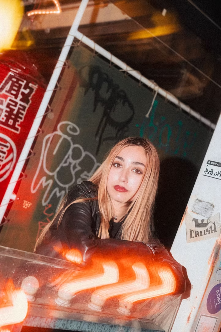 A young woman with long blonde hair and red lipstick leaning on a counter in a bar or club with graffiti and neon signs on the walls behind her.