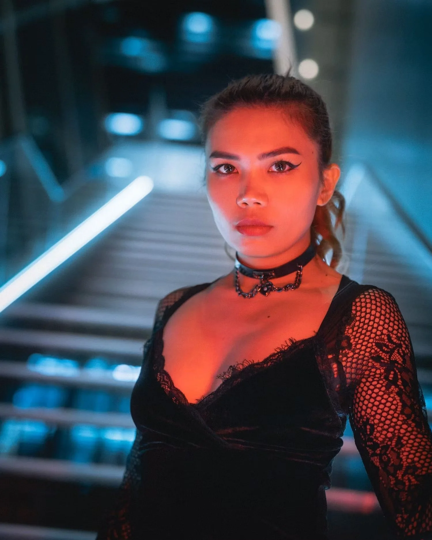 Portrait of a young woman with dark hair styled in a ponytail, wearing black lace top and choker necklace, with makeup including winged eyeliner, standing in front of a blurred background with neon lights.