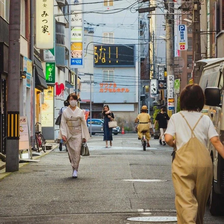 A quiet Tokyo street with pedestrians in casual outfits, lined with small shops, overhead power lines, and Japanese signs. A woman in beige overalls walks ahead.