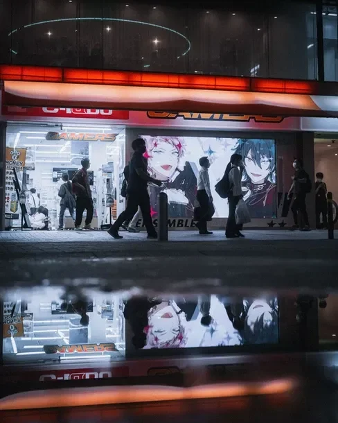 Reflections of pedestrians in puddles, with anime posters glowing. Captured during a Tokyo street photography workshop.