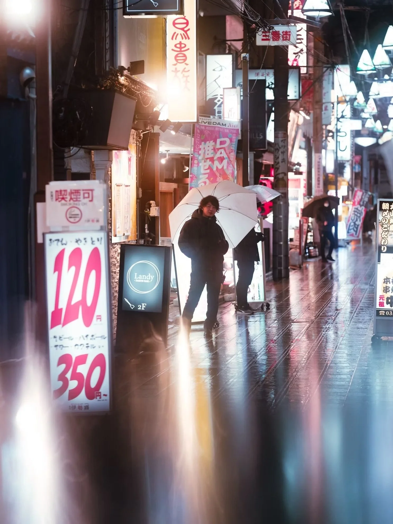 A street scene in Japan at night with bright neon signs, people with umbrellas, and reflections on a wet sidewalk.