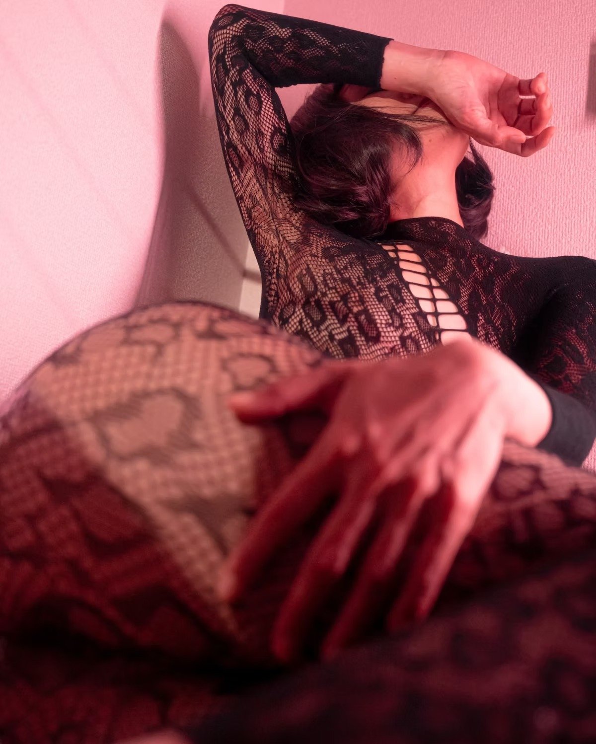 A woman wearing black lace lingerie with snake print, sitting on a bed under pink lighting. Boudoir Photography by Tokyo Photographer Deniz Demir. Japan.