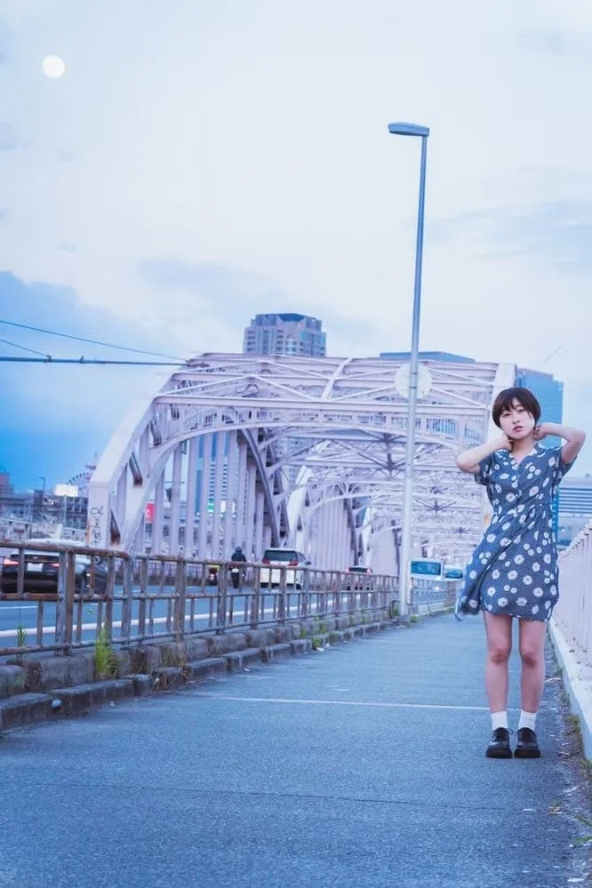 A young woman in a blue polka dot dress standing on a sidewalk near a white bridge, with city buildings in the background and an overcast sky.
