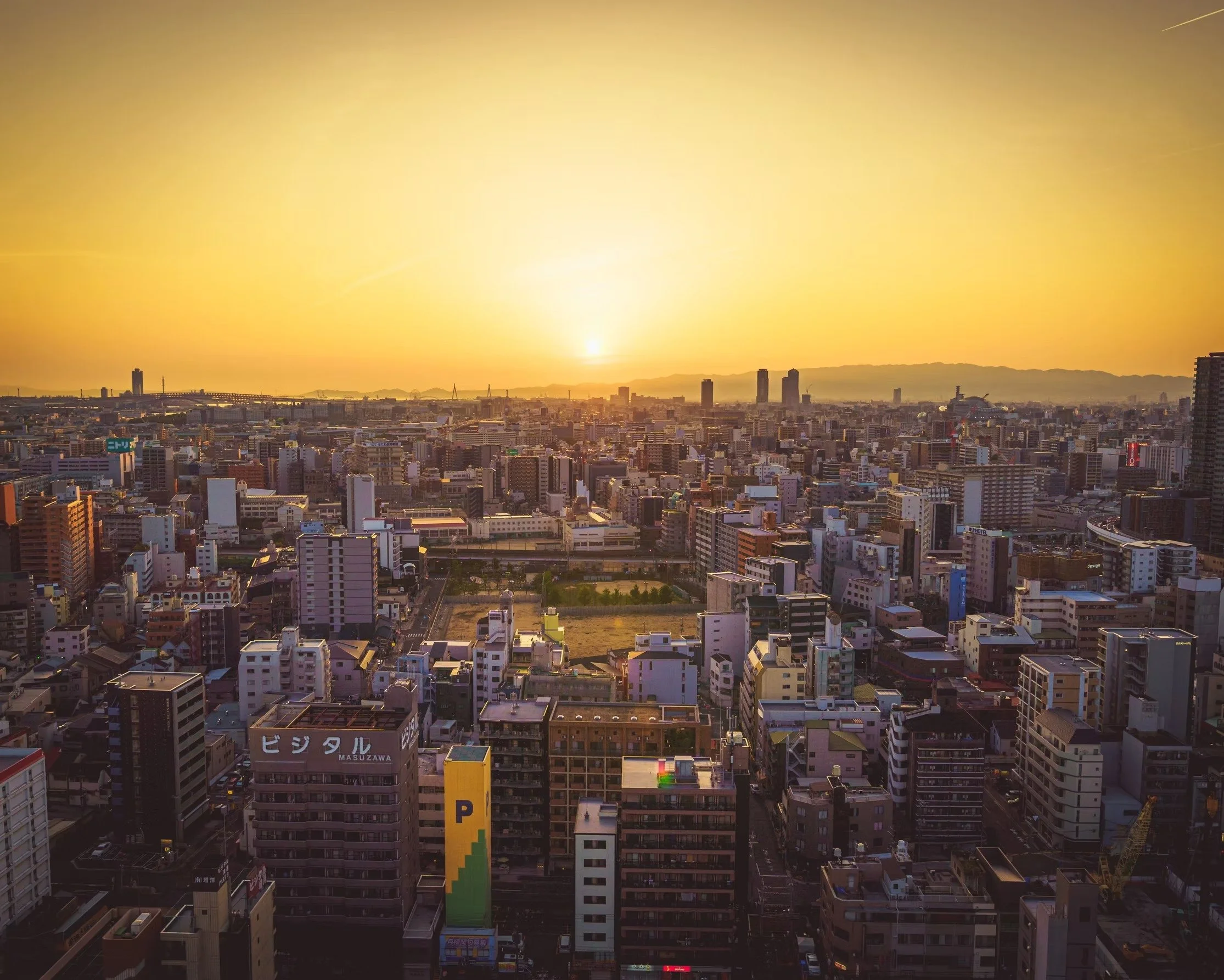 Cityscape at sunset with numerous buildings and skyscrapers, mountains in the distance, and a yellow-orange sky.