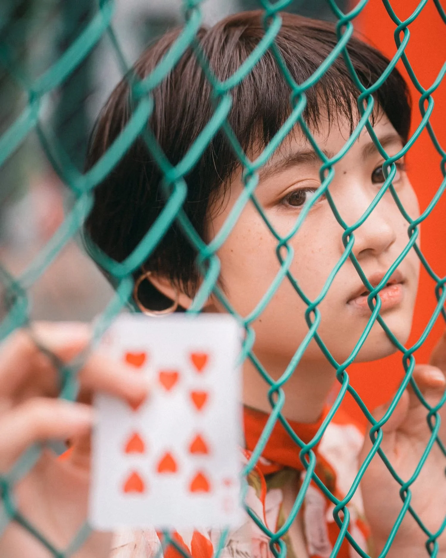 A young girl peering through a green chain-link fence, holding a white card with red heart-shaped cutouts, with a blurred background.