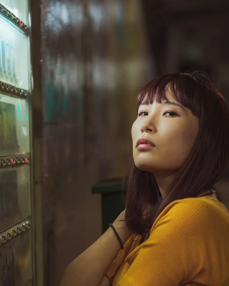 A young woman with dark hair and bangs wearing a yellow top, standing next to a lit vending machine in an indoor setting.