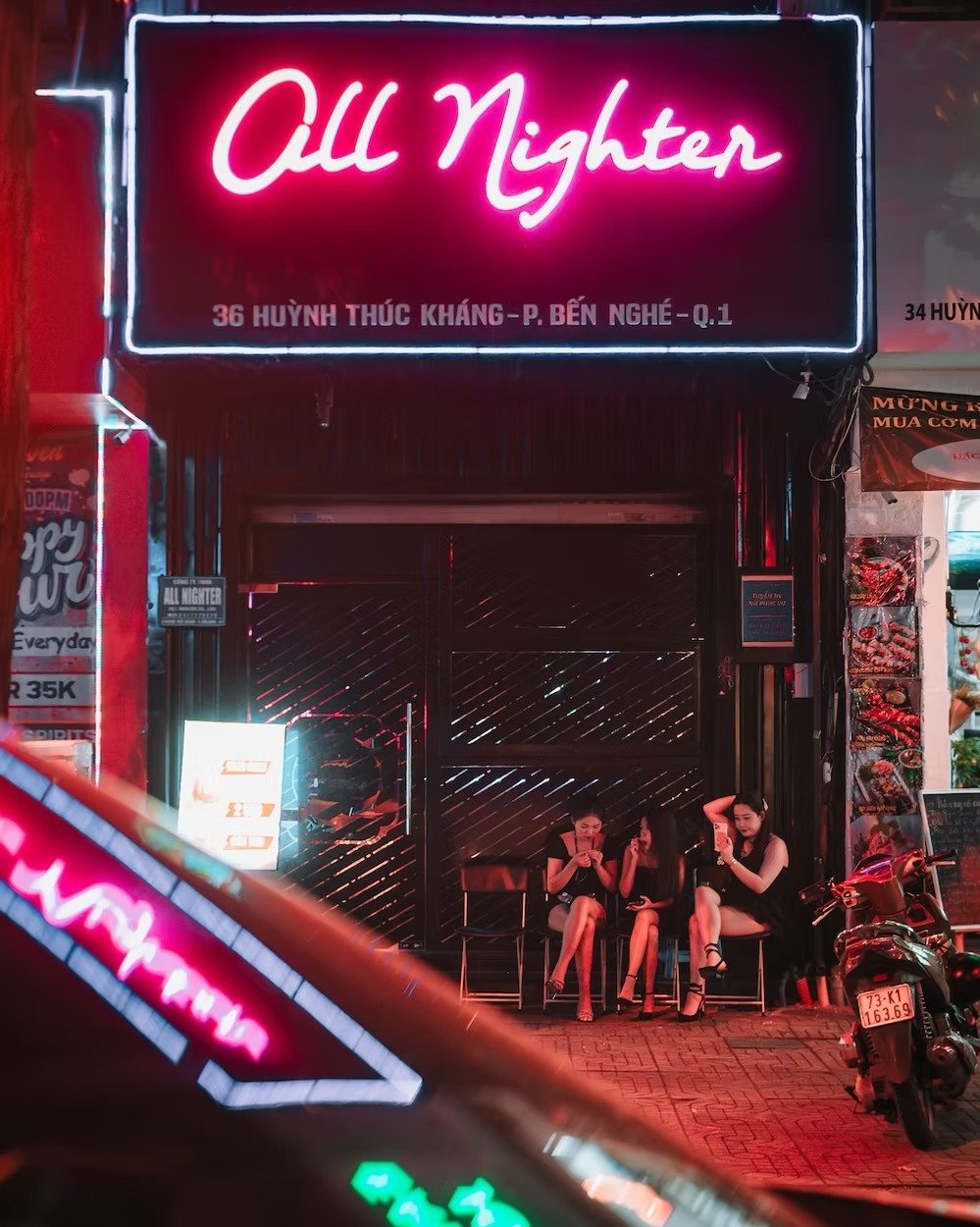A neon sign reads 'Vigil Nighten' above the entrance of a building with three women sitting outside, looking at their phones, illuminated by surrounding neon lights at night. Shot by street photographer Deniz Demir on the streets of Saigon Vietnam.