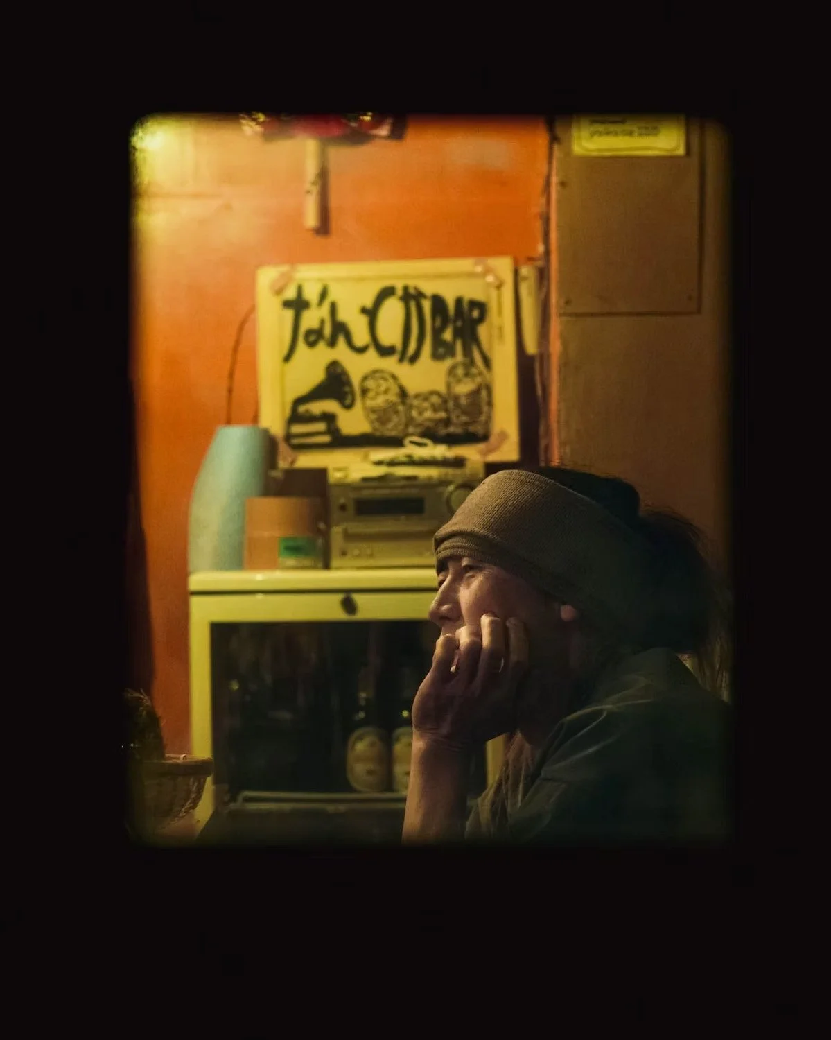 A Japanese man wearing a headband sits in a dimly lit room with her hand on her face, looking contemplative. The background features a sign that reads 'John CUBA' with a drawing of two barrels and a shoe, along with a stereo and a refrigerator. Captu