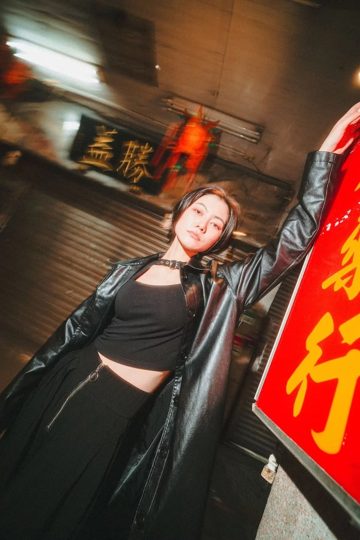 A young woman with short dark hair posing with one arm raised and a confident expression, standing in a street at night with Taiwanese signs and illuminated storefronts in the background.