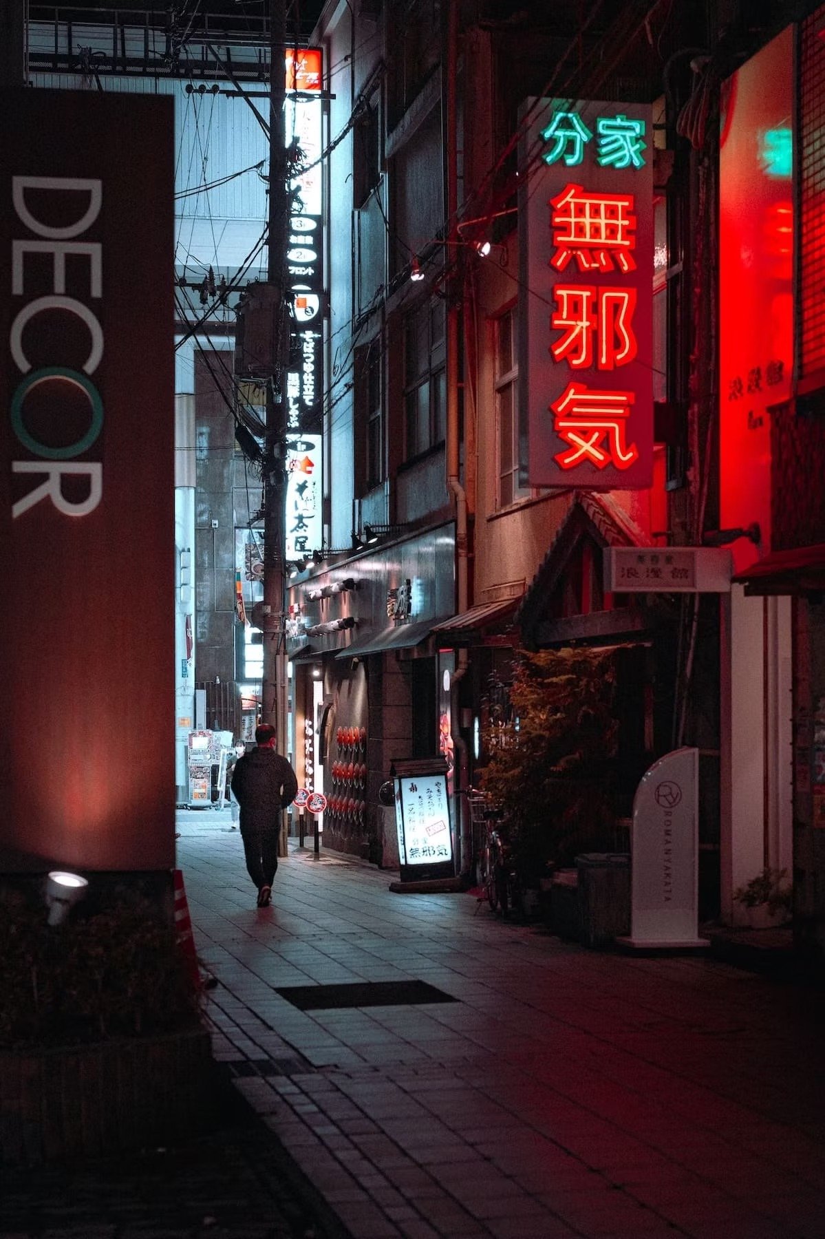 A narrow urban street at night illuminated by neon signs in Japanese characters. A person is walking away from the camera along the sidewalk, with buildings, wires, and additional illuminated signs in the background. Kagoshima shot by Tokyo street ph