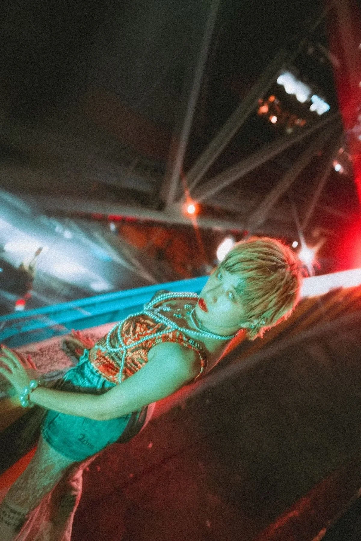 A young person with short, blonde hair and colorful makeup, wearing a patterned sleeveless top, is lying on a red ledge in a neon-lit environment, possibly a nightclub or concert venue in Osaka.