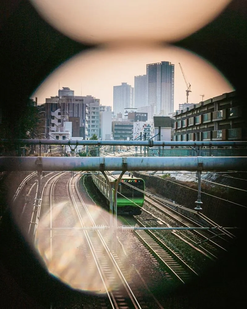 A green train on railway tracks viewed through a circular peephole with an urban city skyline in the background.