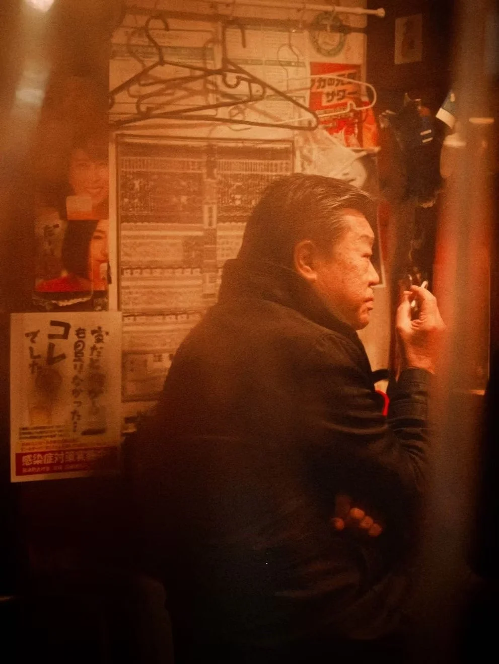 A man sitting in a dimly lit room, smoking a cigarette, with posters and hangers on the wall behind him.