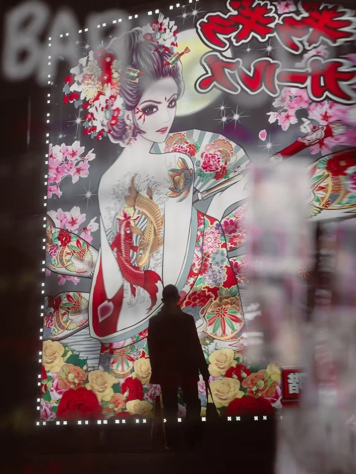 Silhouette of an elderly person standing in front of a large, colorful wall mural featuring a woman with traditional geisha makeup, elaborate hair decorations, and kimono with floral and fish designs, set against a moonlit background with cherry blossoms and flowers.
