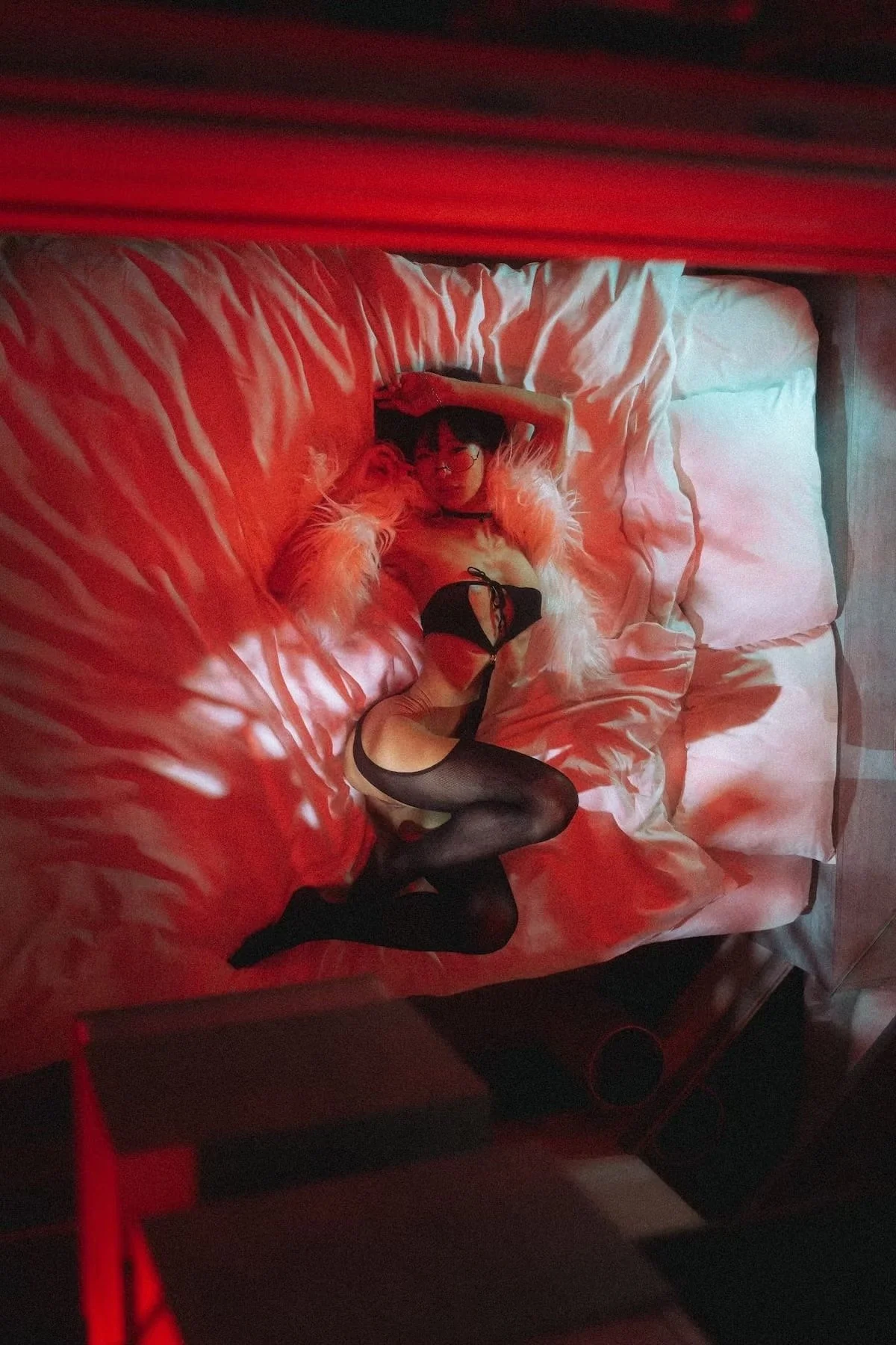 A woman with long pink hair, wearing black lingerie, fishnet stockings, and glasses, lying on a bed with white sheets and pillows, illuminated by red lighting.