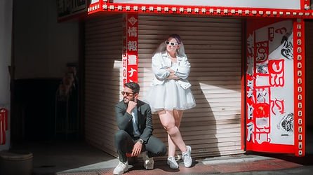 A man and woman posing outside a closed shop with red and white signage, the woman standing with arms crossed and the man squatting with hand on chin.