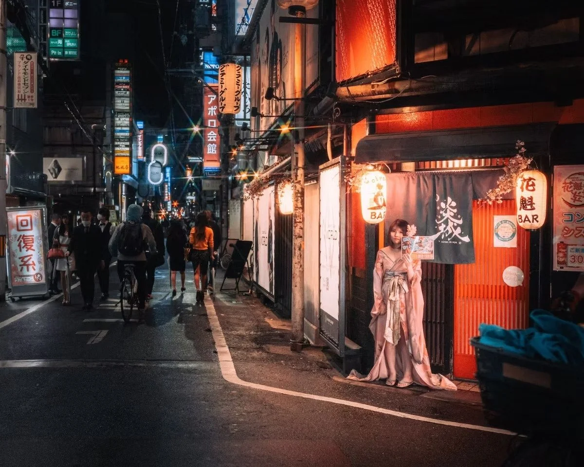 Nighttime street scene in Japan with illuminated signs, pedestrians, and a woman in kimono taking a selfie outside a bar shot by Tokyo street photographer Deniz Demir.