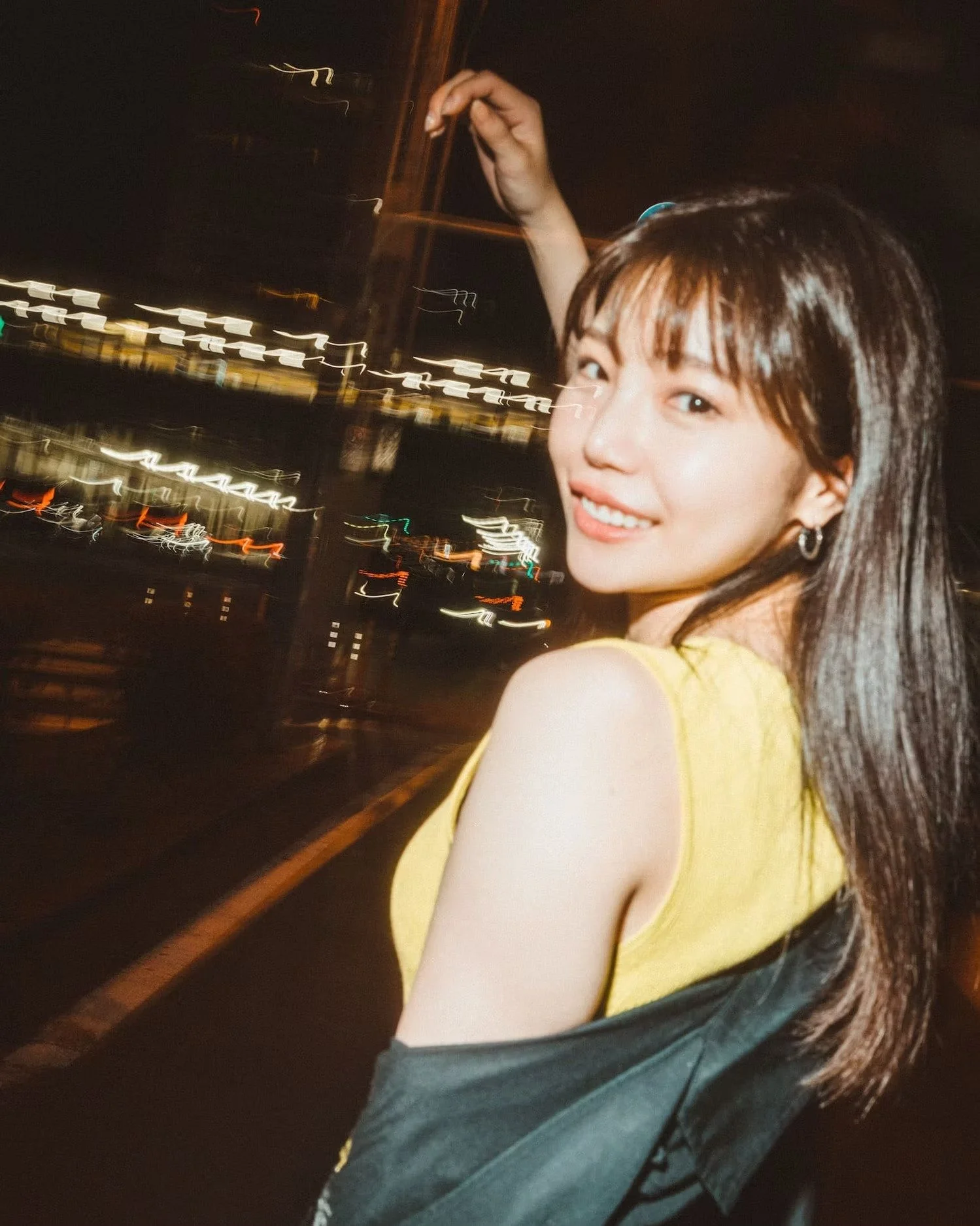 A young woman with long dark hair and a yellow top, smiling at night with city lights in the background.