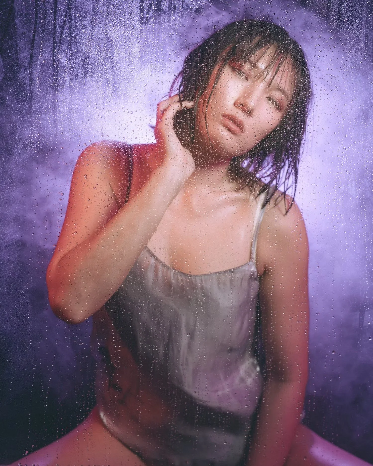 A young woman with wet hair, posing behind a rain-covered glass with purple and blue lighting.