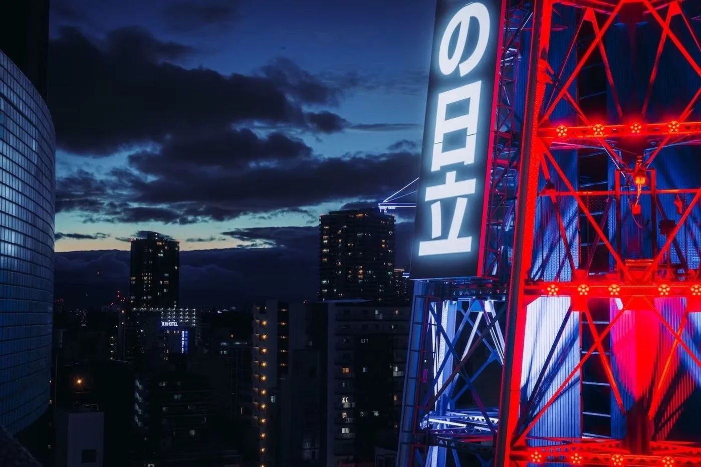Nighttime cityscape with illuminated buildings and a large neon sign in Japanese characters on a red and blue metal tower.