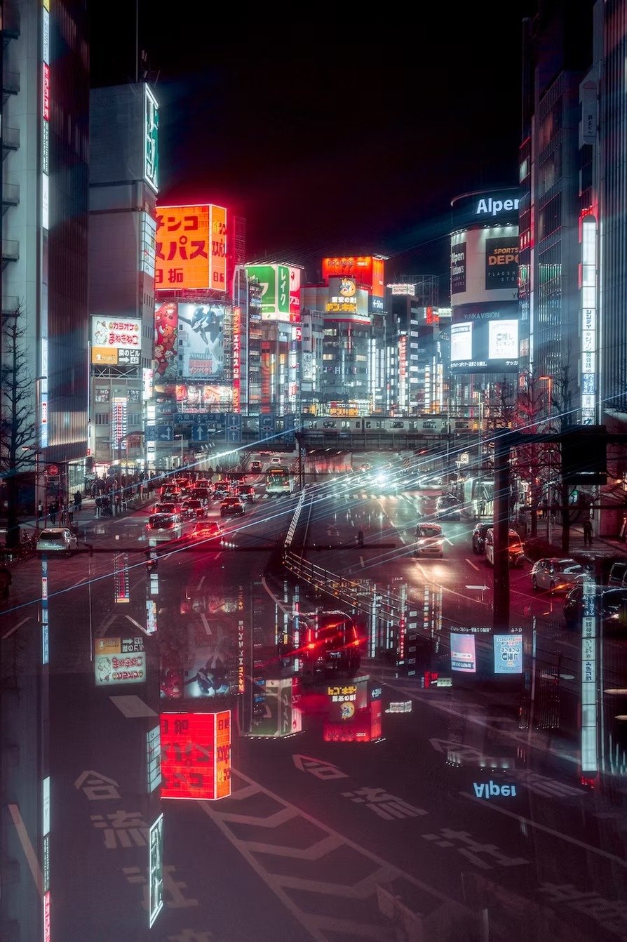 Nighttime city street scene in Japan with bright neon signs, tall buildings, and cars on the road, reflecting city lights.