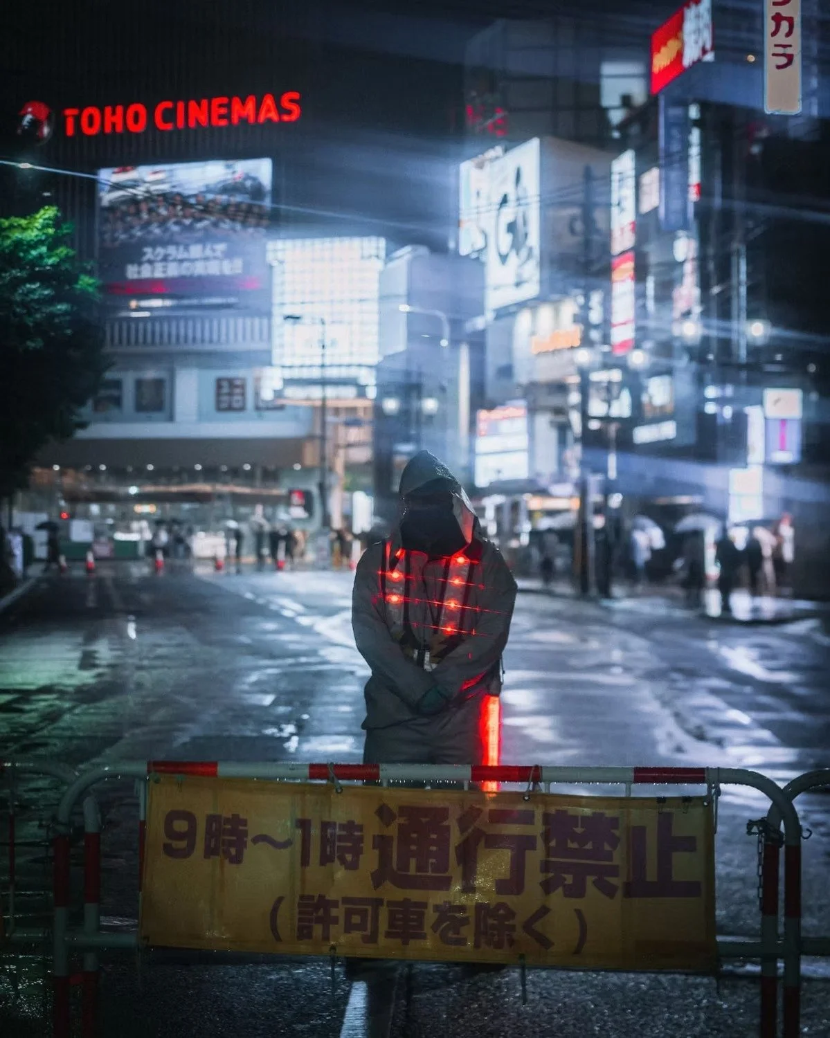 A person wearing a hoodie with glowing red lights standing behind a barrier with a Japanese warning sign in a city street at night. The street is wet and illuminated by numerous bright billboards and neon signs. Street photography by Deniz Demir.