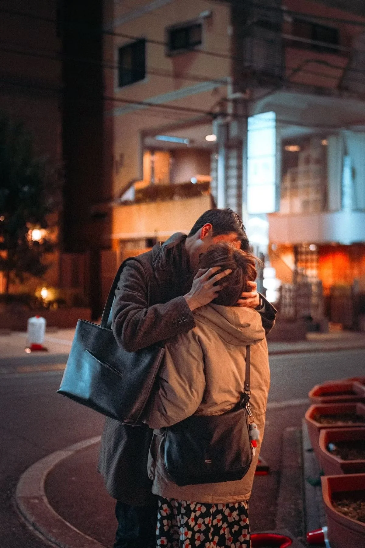 Two people embrace on a city street at night, one with a black tote bag and dark jacket, the other with a beige jacket and patterned skirt, against a background of illuminated buildings and street lights. On the streets of Ikebukuro.