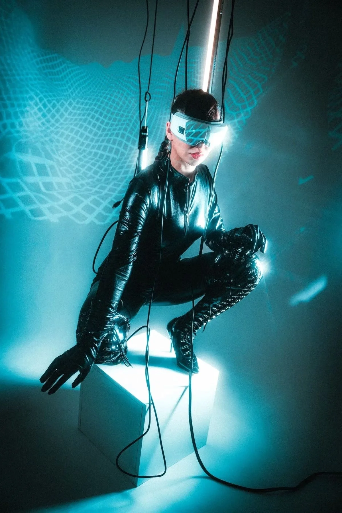 A woman wearing futuristic-looking virtual reality goggles, a black leather jacket, and matching leather gloves and boots. She is crouching on a white cube illuminated from below, with a dark background featuring a digital grid pattern and glowing bl