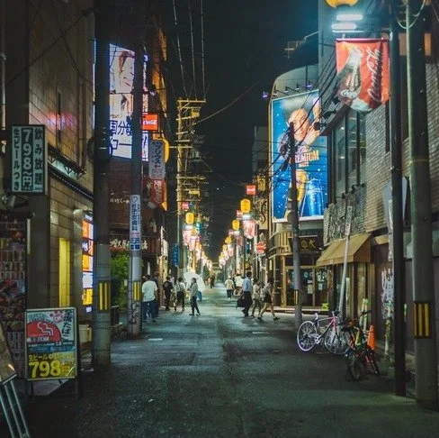 A narrow Tokyo alley glows with neon signs and lanterns, as bicycles rest near small shops and distant pedestrians walk under electric wires.