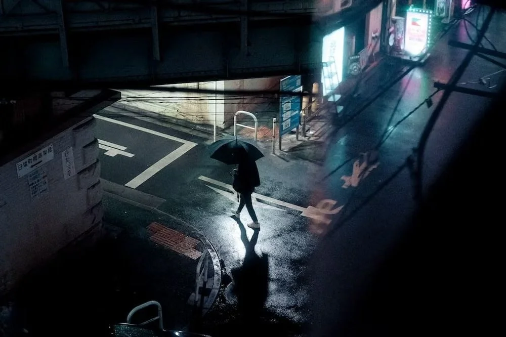 A person walking with an umbrella on a rainy night in an urban area, illuminated by streetlights and neon signs, with reflections on the wet pavement. Shimbashi night scenes.