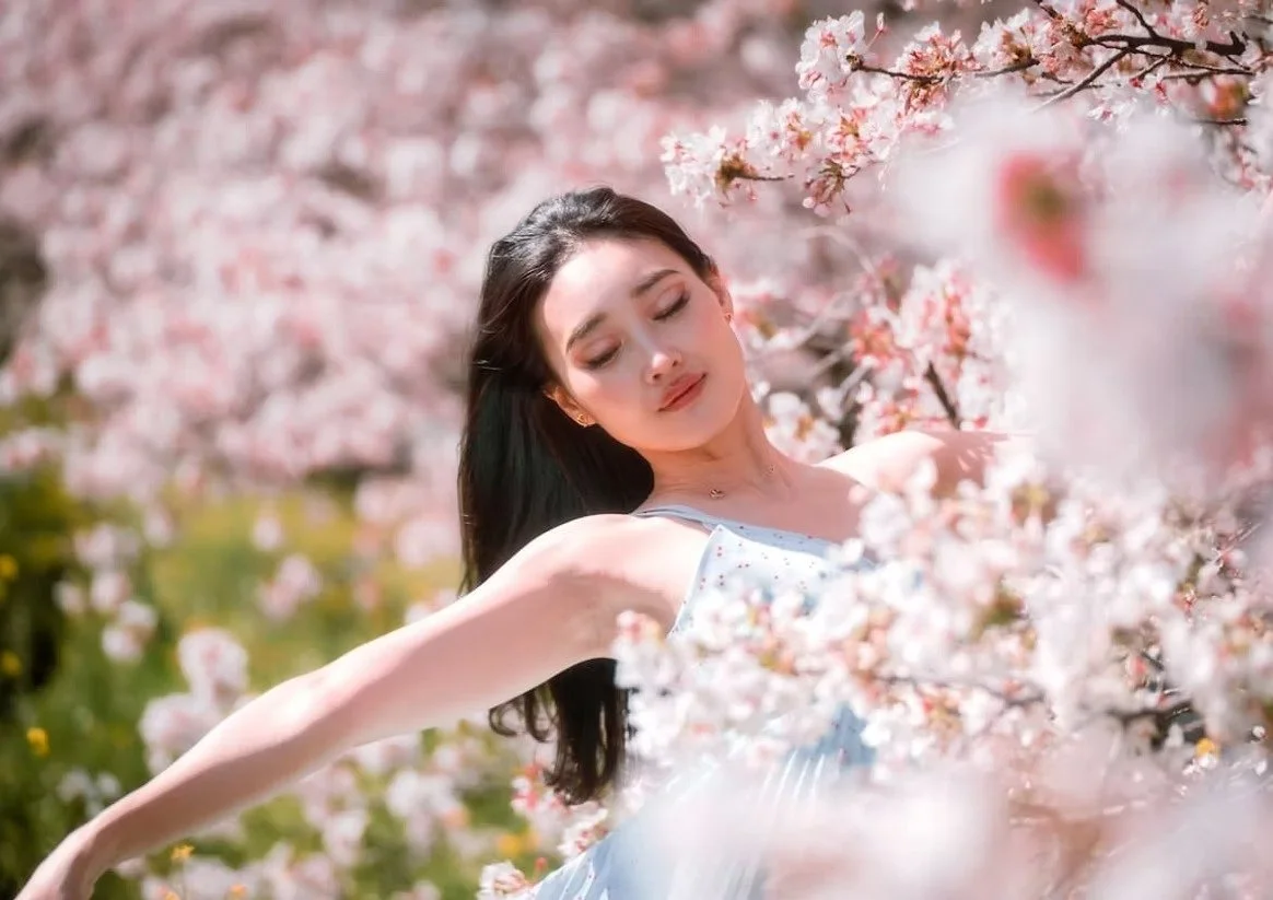 A woman in a light blue dress closes her eyes and dances gracefully among the cherry blossoms. The pink flowers frame her as she enjoys the serene moment, with sunlight filtering through the blossoms.