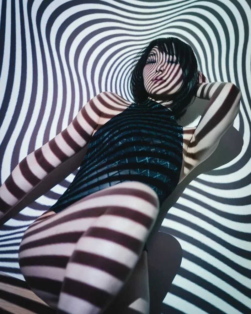 A woman with short black hair posing against a background with black and white wavy stripes projected onto her and the surroundings.
