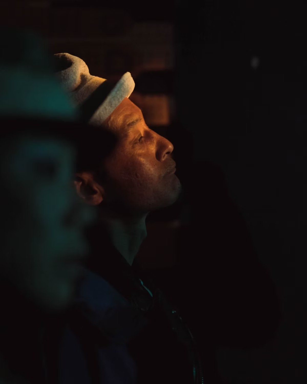 Side profile of a Japanese man in Koenji with glowing skin illuminated by warm light, looking upward, with a dark background.