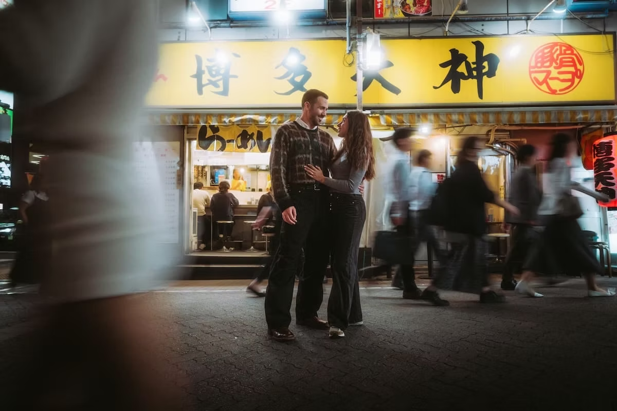 A couple stands close together, smiling and looking at each other on a city sidewalk at night, with a Japanese restaurant in the background and blurred pedestrians walking by.