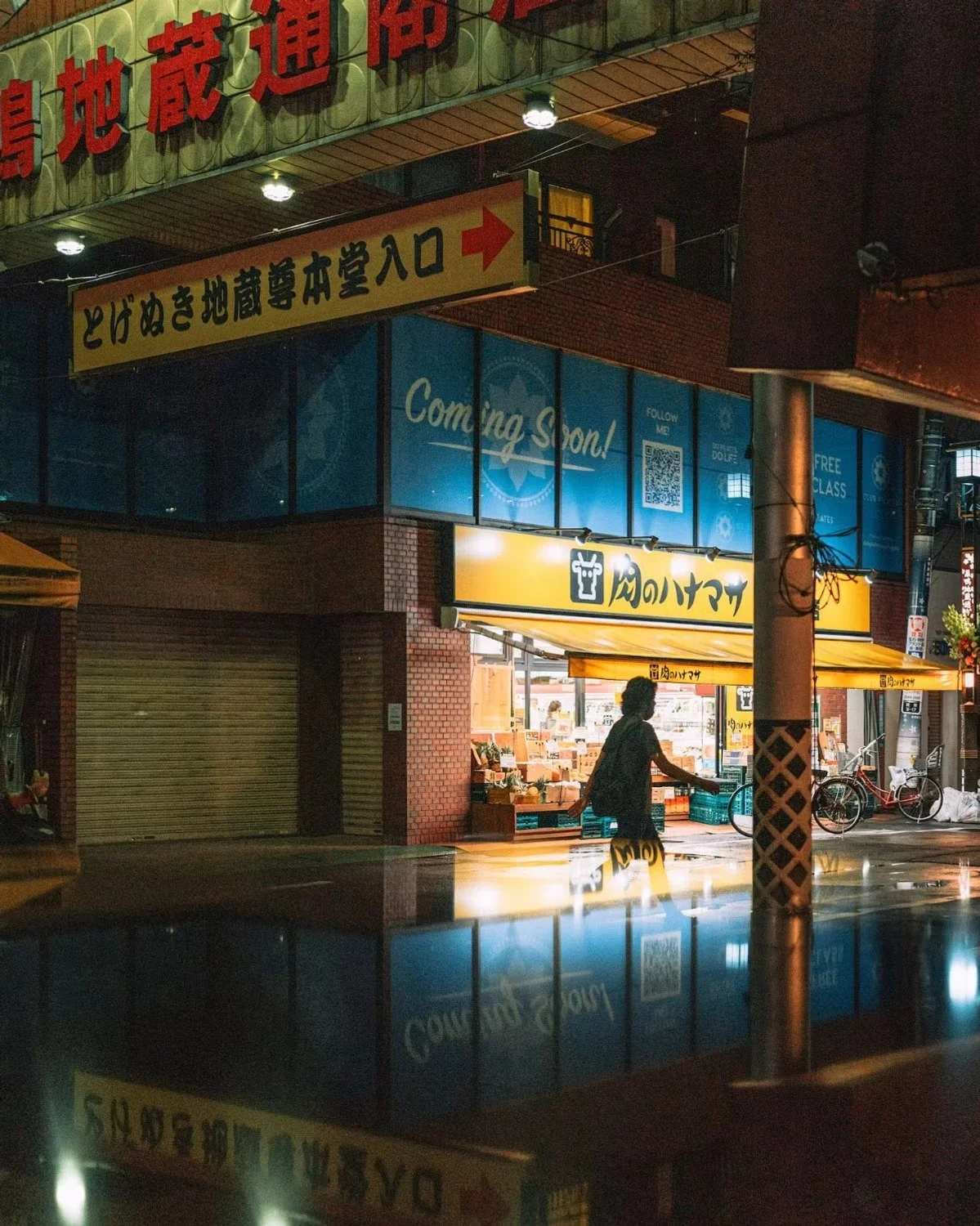 Nighttime street scene with a person walking past a store with bright signs in Japanese. The store has yellow signs and is lit from within. Bicycle bikes are parked outside, and the street is wet, reflecting the lights.