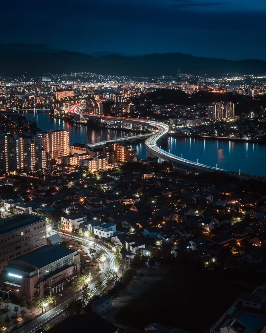 Night cityscape with illuminated roads, buildings, and a river, with mountains in the background.
