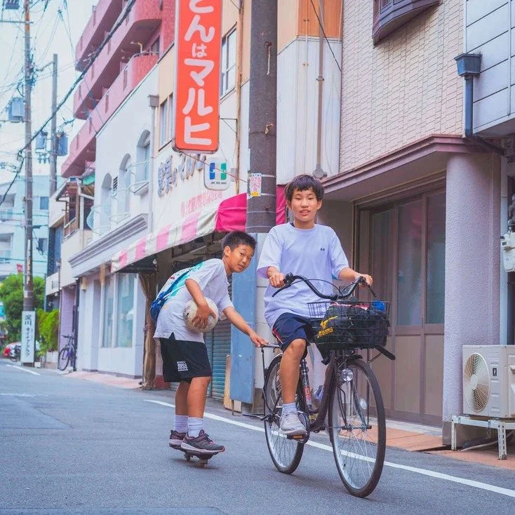 Two kids, one on a bicycle and the other on a skateboard, riding down an Osaka street, captured during slow travel.