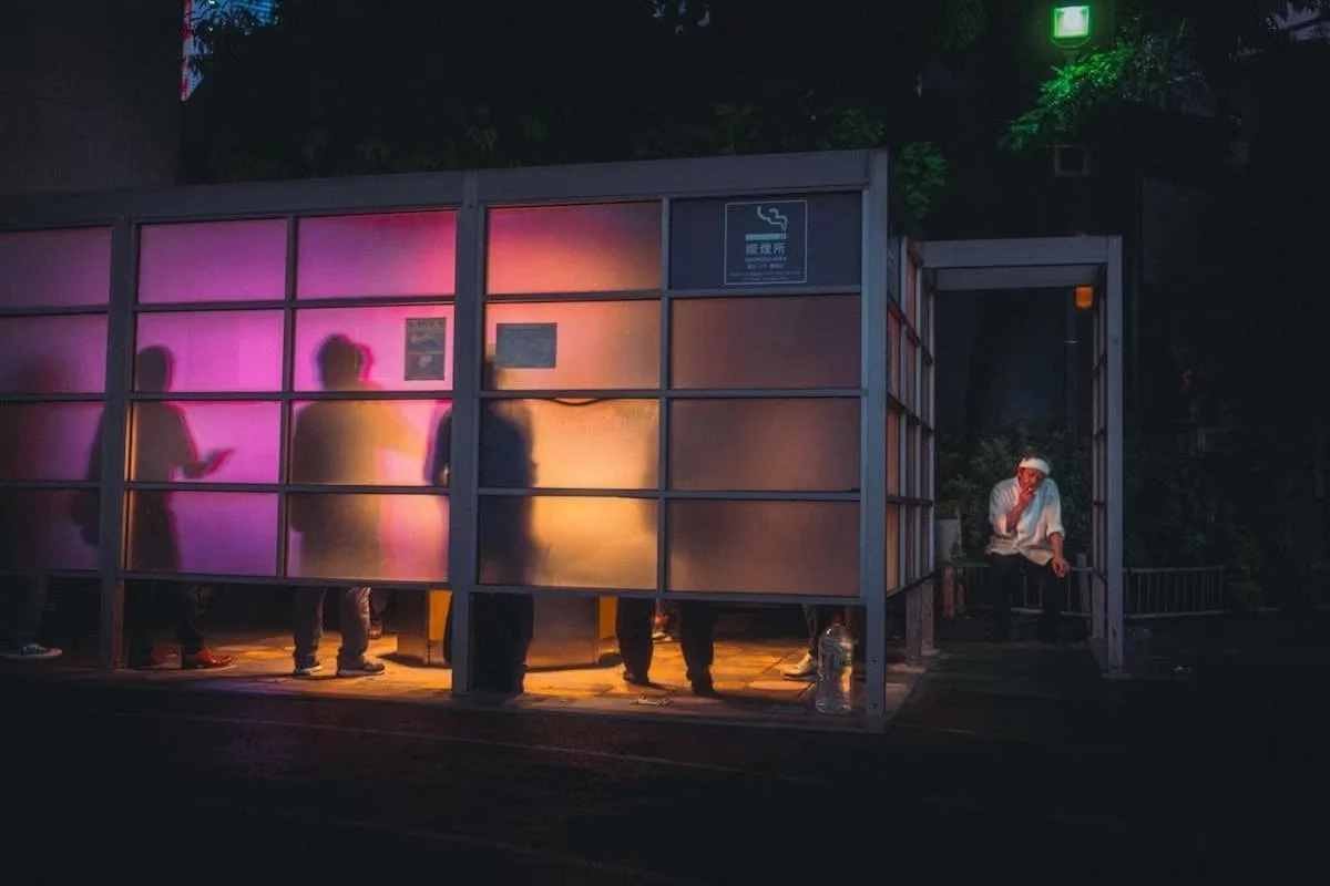 Silhouettes of people standing inside a bus stop with frosted glass panels, illuminated by pink and orange lights, and a seated person wearing a bandana outside the bus stop at night.Tokyo street photography by Deniz Demir.