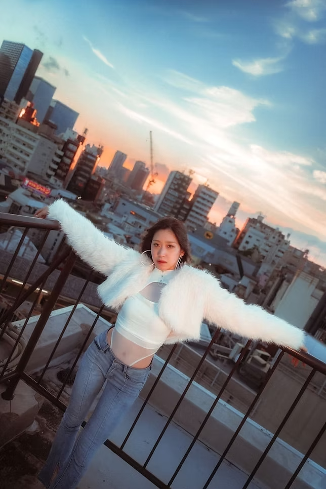 Young woman standing on a rooftop balcony with city buildings in the background during sunset, wearing a white crop top, light blue jeans, and a fluffy white jacket.