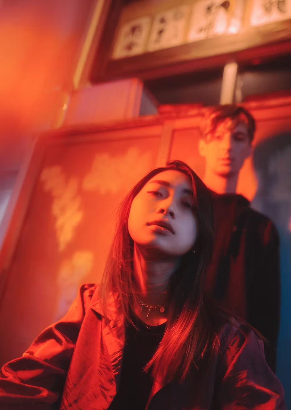 A young woman with long hair and a choker necklace looks into the camera, illuminated by red lighting, with a young man with glasses standing behind her in the background, inside a dimly lit room with framed pictures on the wall.