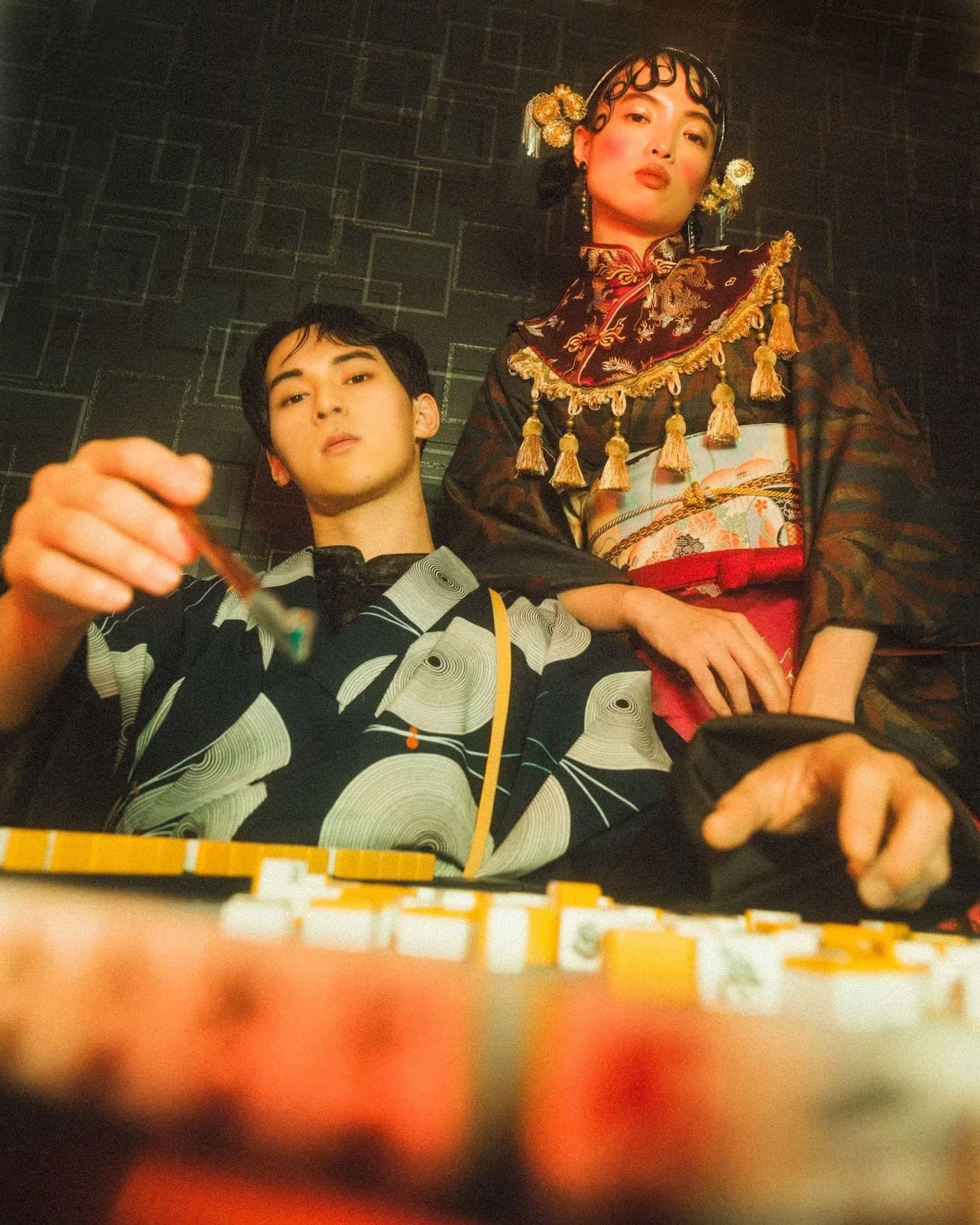 Two young people playing a game of poker, with poker chips on the table, in a dimly lit room with dark tiled walls. The person on the left, wearing a black and white patterned shirt, reaches with their hand, while the person on the right is dressed i