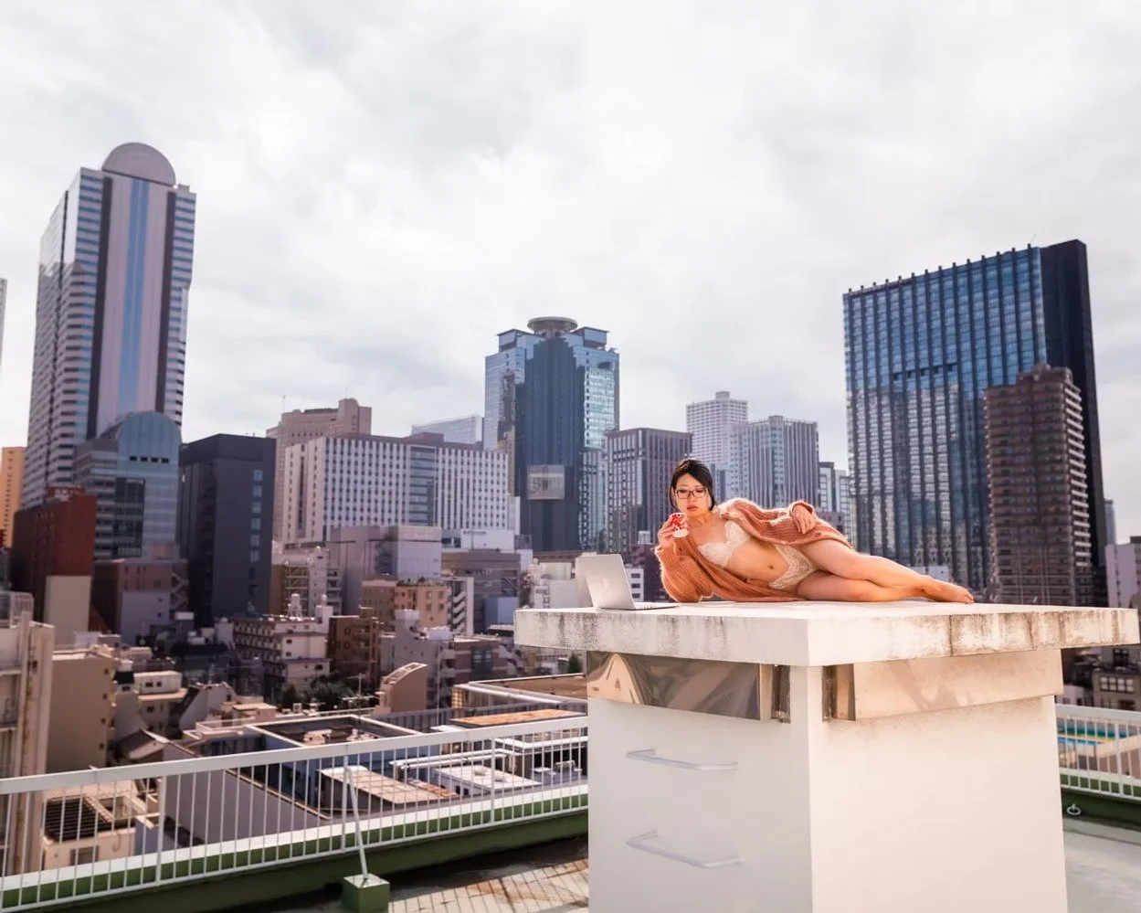 A woman in lingerie and a brown coat lying on a rooftop ledge with a city skyline in the background in Shinjuku shot by Tokyo portrait photographer Deniz Demir.