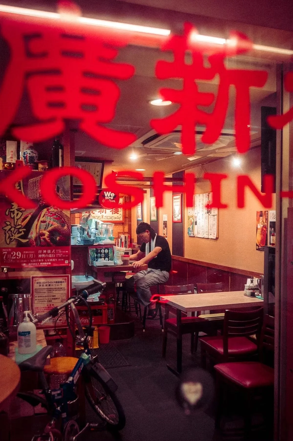 View through restaurant window with red lettering. Inside, a man is sitting at a table, wearing glasses and a black shirt with a white towel around his neck. There are chairs, posters, and decor on the walls, and various items such as a rice cooker a