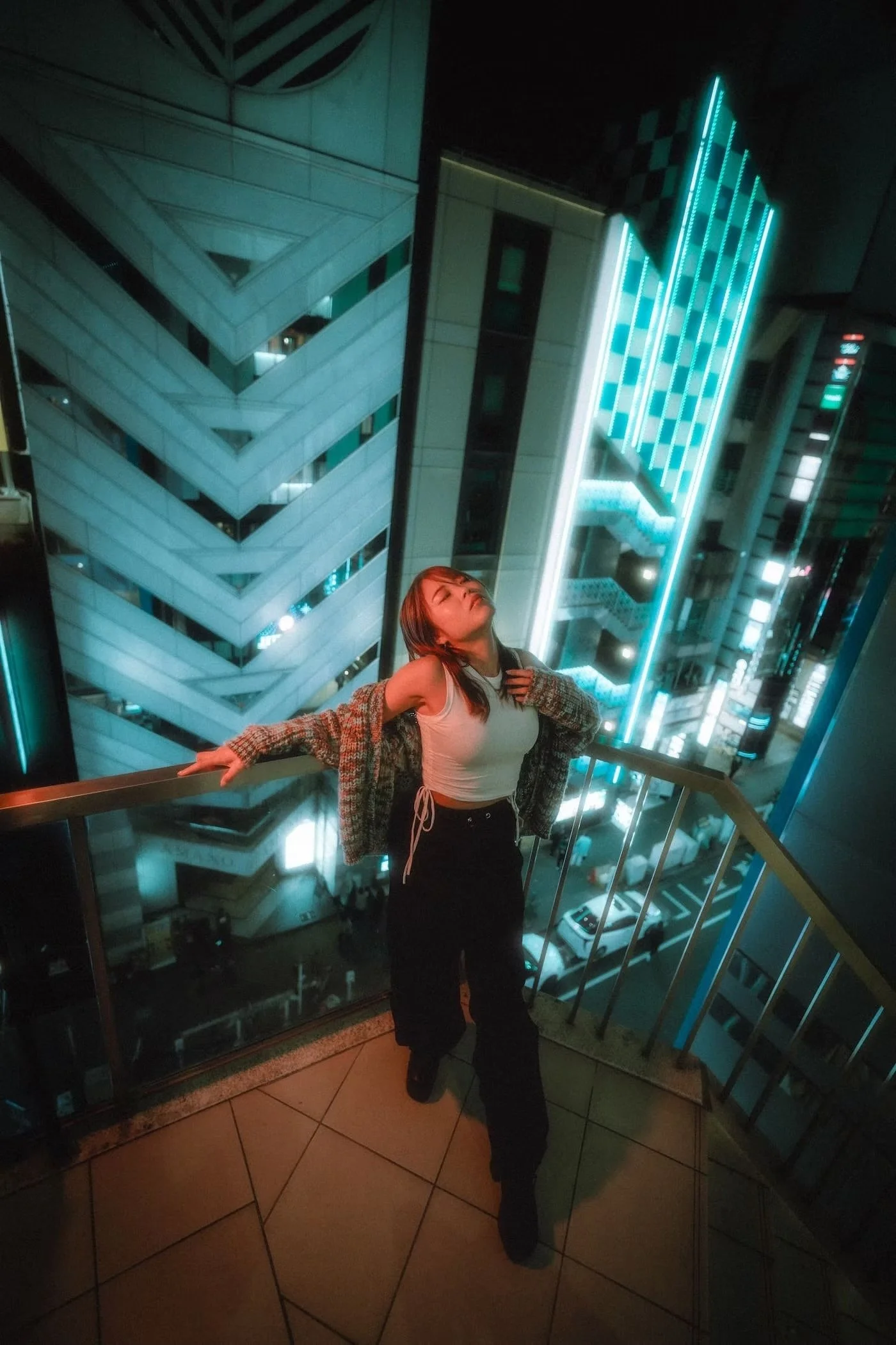Young woman with long hair, wearing a white cropped top and loose black pants, posing with arms outstretched on a balcony at night with city buildings illuminated in blue neon lights in the background.