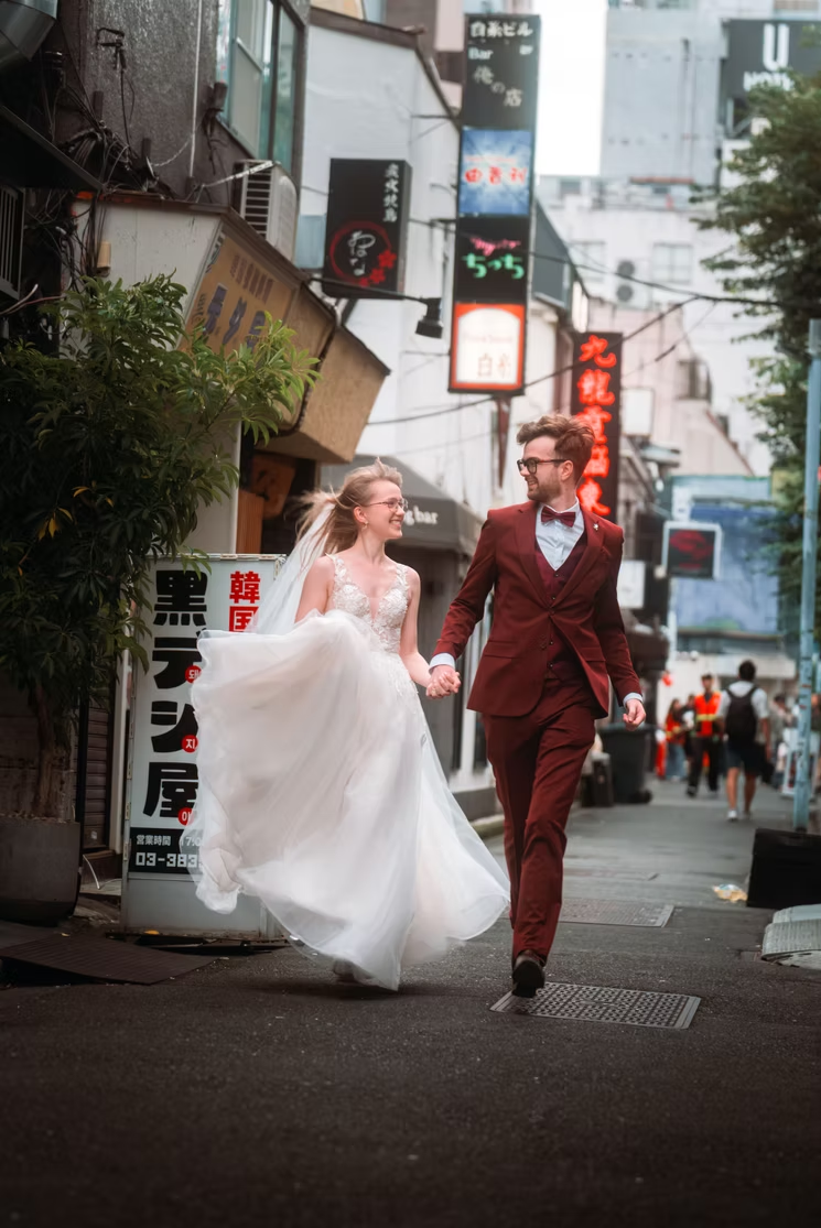 A newlywed couple walking hand in hand on a city street with Asian signage, the bride in a white wedding gown and the groom in a burgundy suit, smiling at each other.