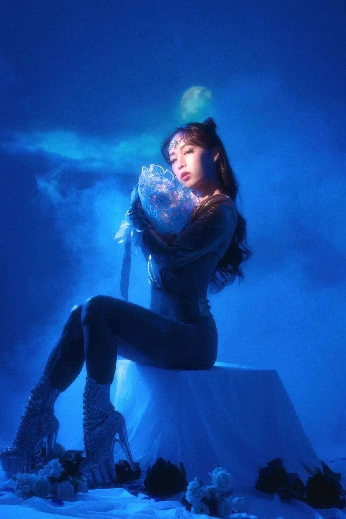 A woman in a dark leather outfit posing with a transparent bouquet, sitting on a white platform surrounded by flowers, with a moon in the background surrounded by clouds.