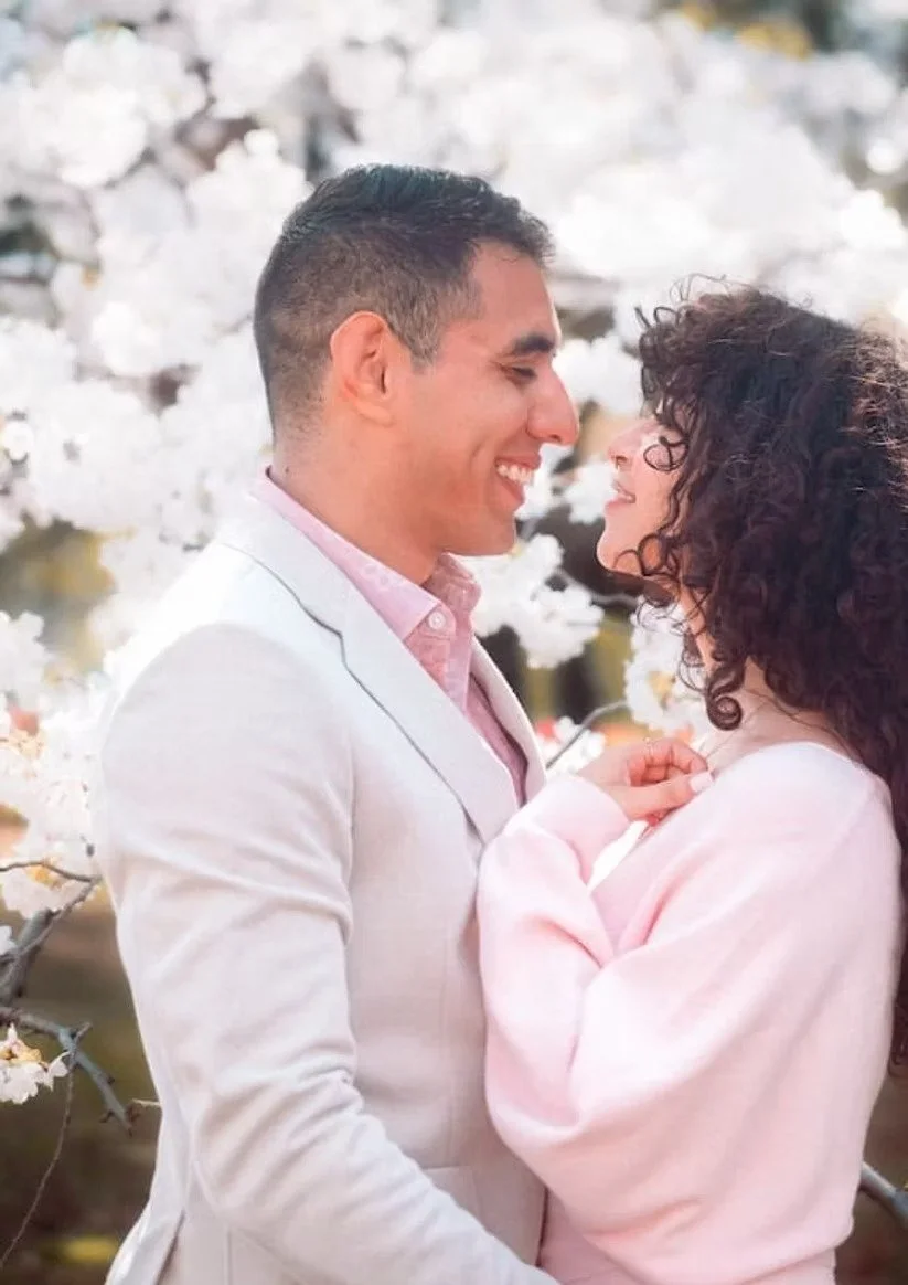 A couple stands together in a cherry blossom garden, sharing a tender moment. The man in a light pink shirt and the woman in a pink sweater smile at each other, with the beautiful cherry blossoms in full bloom surrounding them.